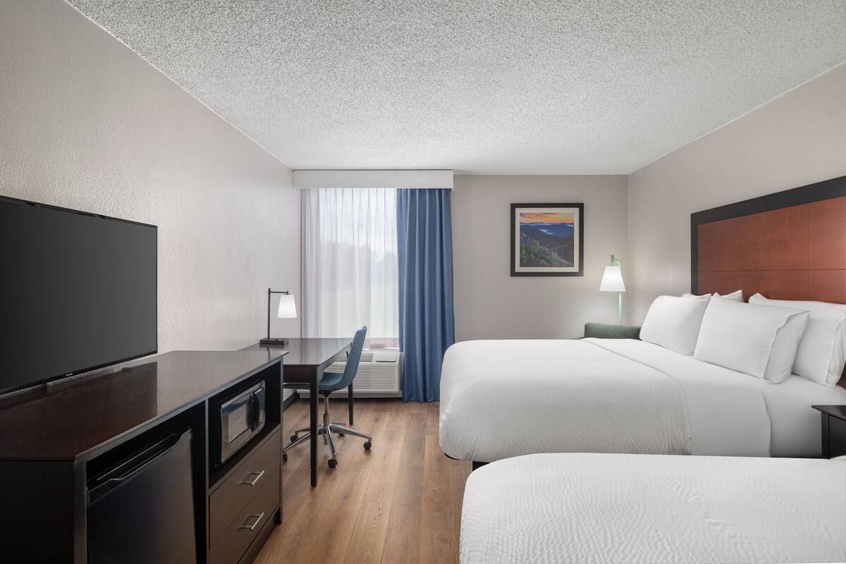 A hotel room with a flat-screen TV, a desk with a chair, two beds, a window with curtains, and a painting on the wall.