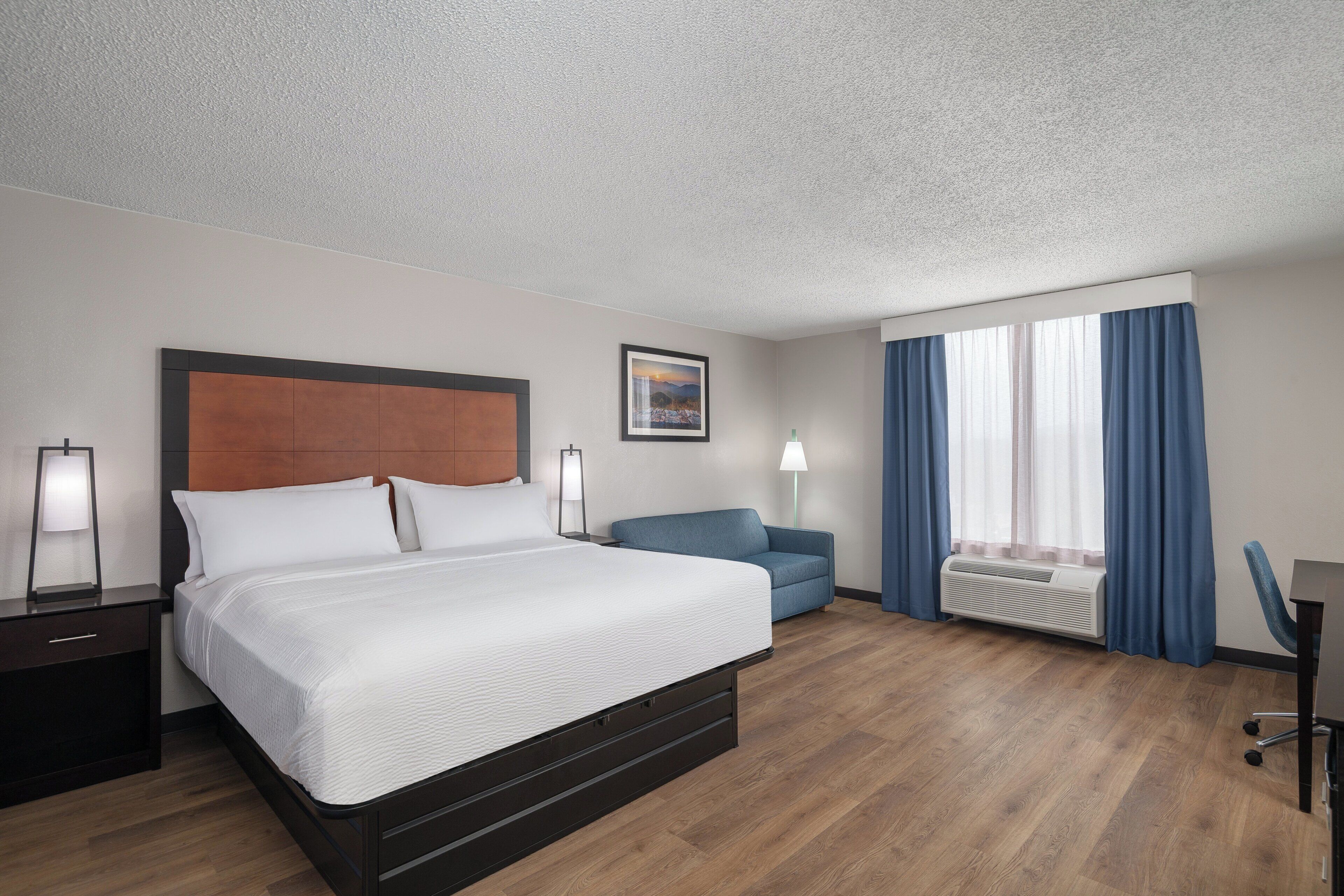 A hotel room with a bed, bedside tables, a desk, a chair, a window with curtains, and a wall-mounted air conditioner.