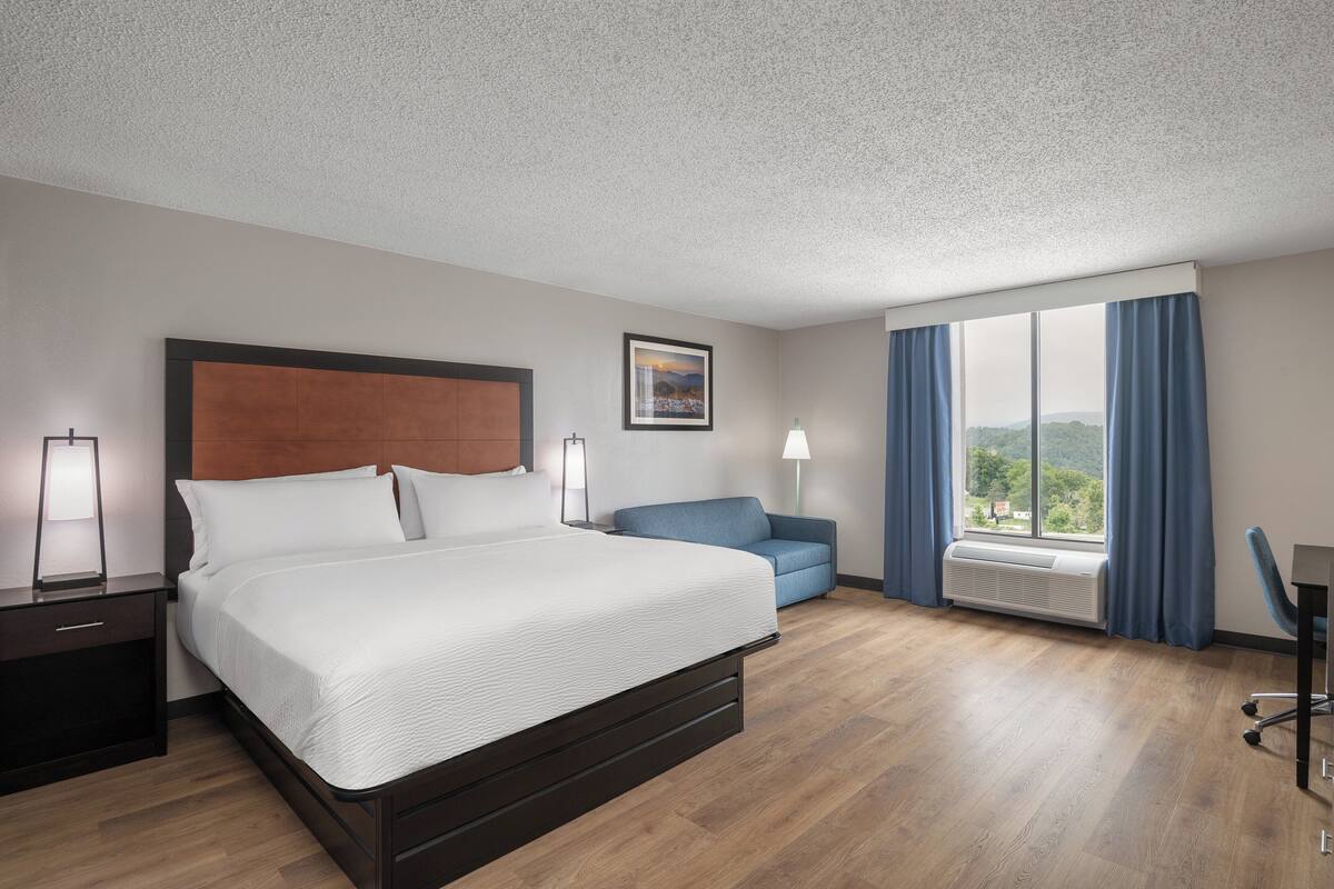 A hotel room with a bed, a desk, a chair, a window with curtains, and a wall-mounted air conditioner.