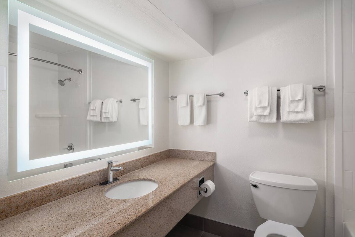 A modern bathroom with a granite countertop sink, a large mirror, a toilet, and multiple towels hanging on a rack.