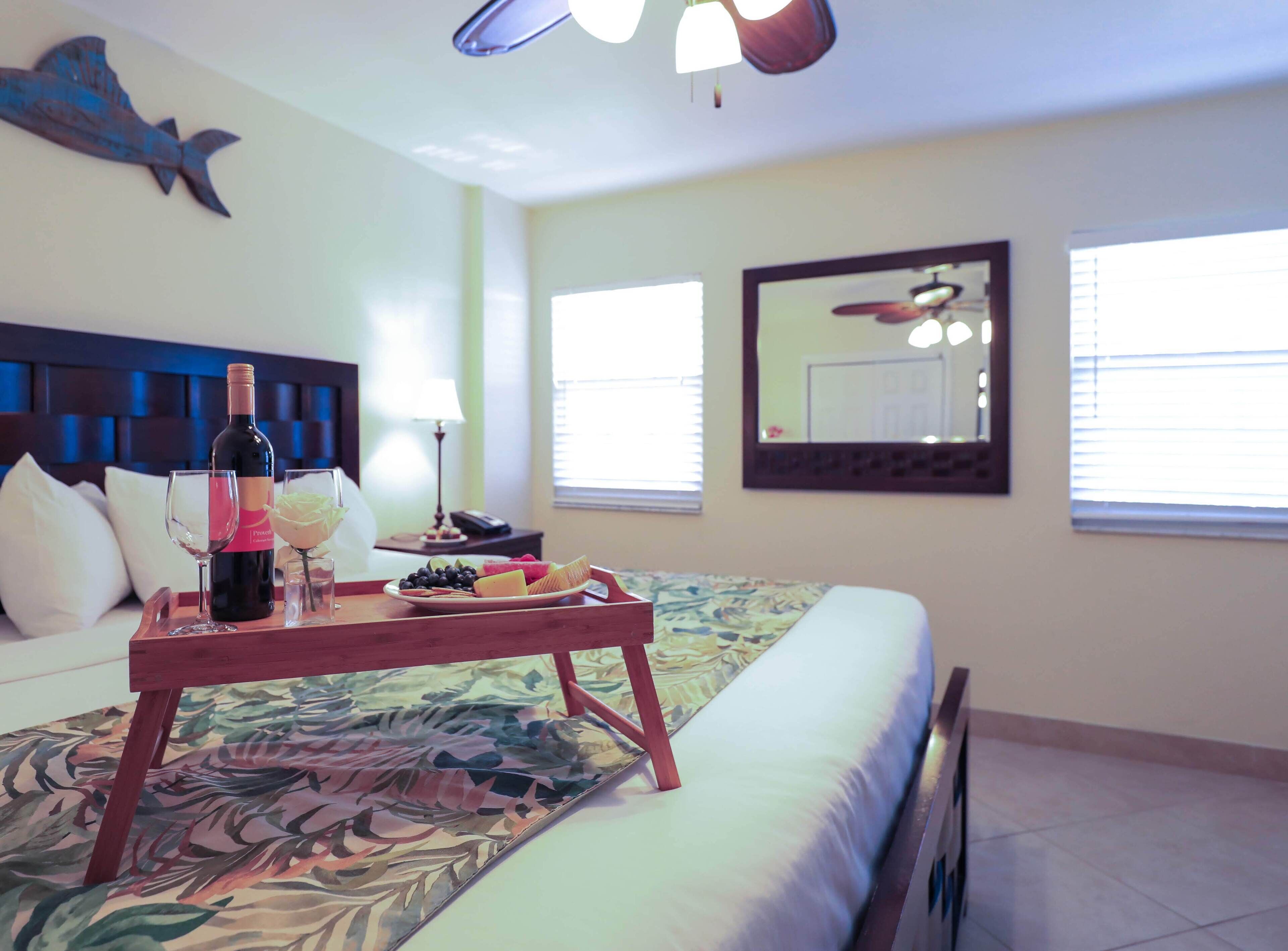 A hotel room with a bed, a bedside table with a tray of food and drinks, a lamp, a ceiling fan, and a large mirror on the wall.