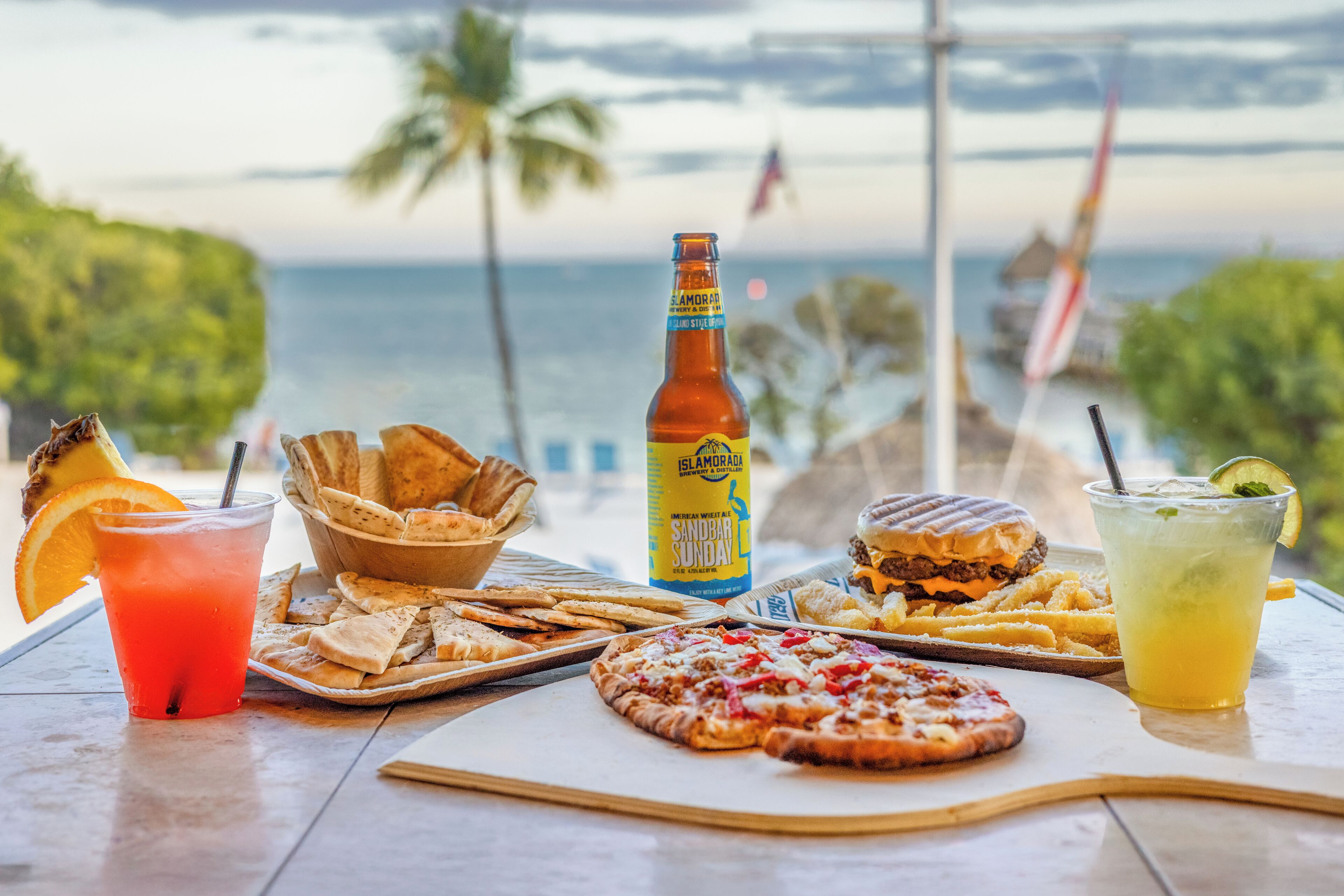 Ein Tisch mit verschiedenen Speisen, darunter ein Burger, Pizza und Pommes, sowie Getränke und eine Bierflasche, vor einer malerischen Meereslandschaft.