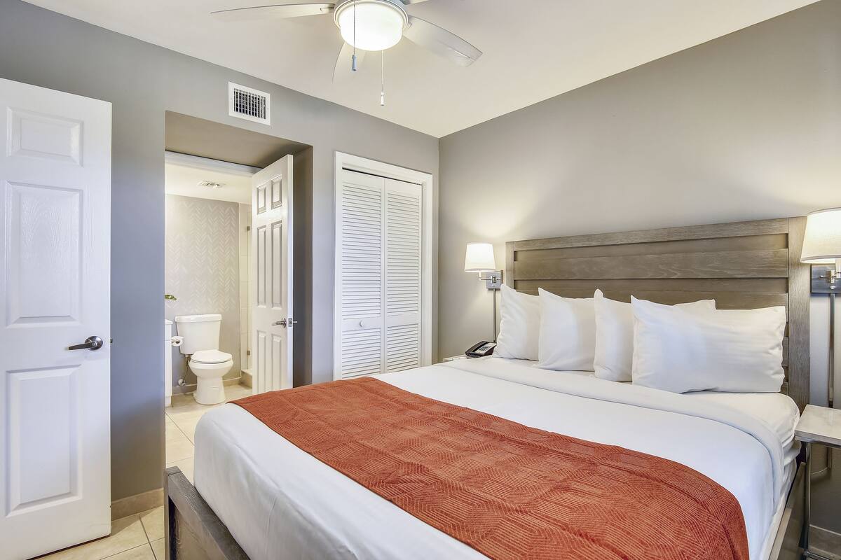 A hotel room with a bed, a nightstand, a lamp, a ceiling fan, and a bathroom visible through an open door.