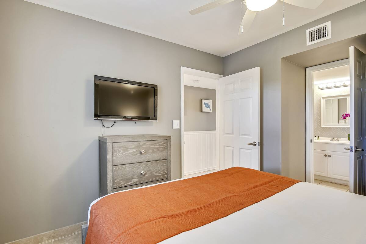 A bedroom with a bed, a television mounted on the wall, a dresser, and a door leading to a bathroom.