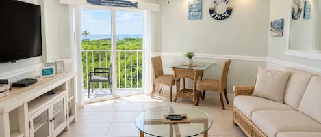 A living room with a television, a glass coffee table, a sofa, and a dining area with wicker chairs.