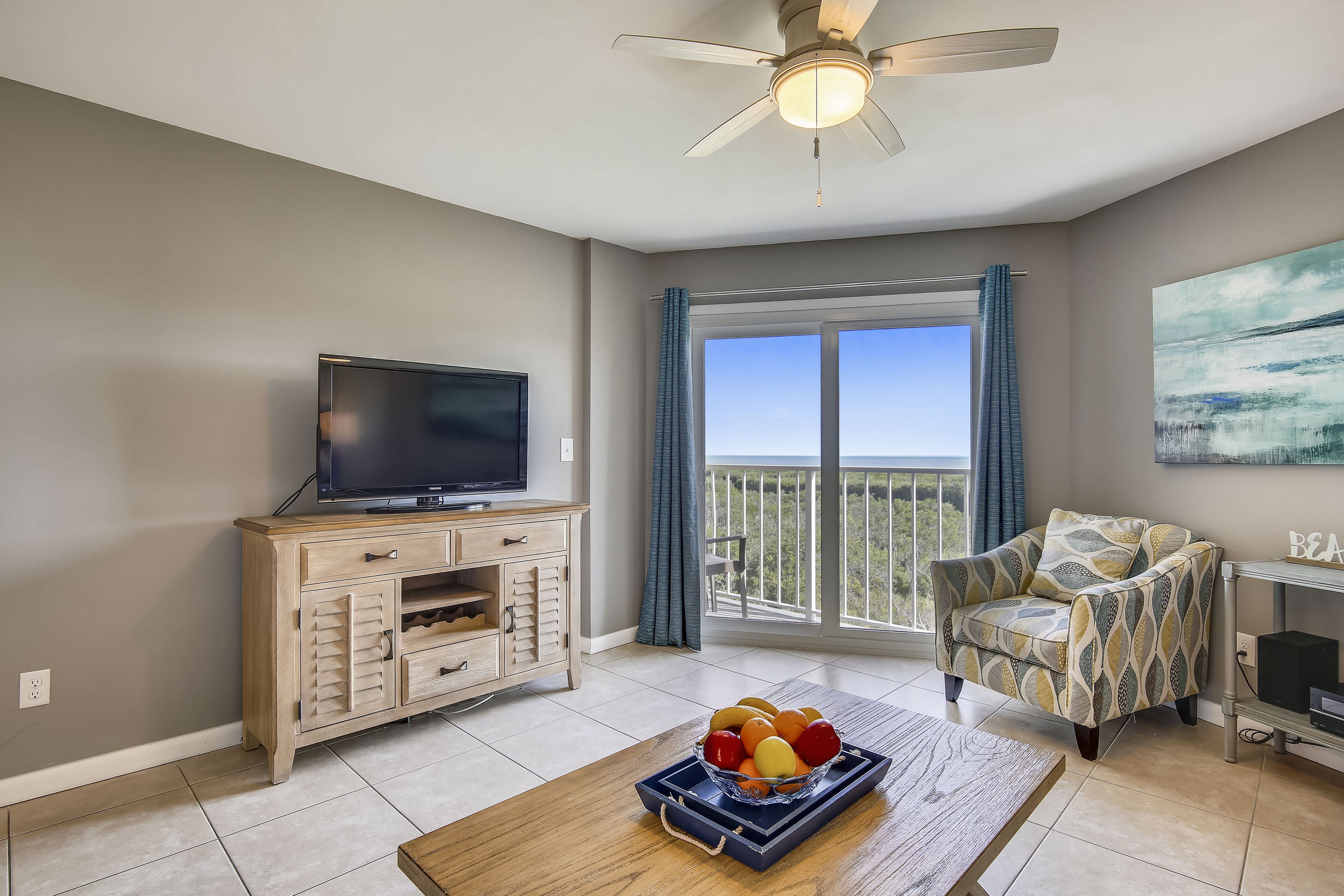 A living room with a television, a wooden cabinet, a patterned armchair, and a balcony with a view.