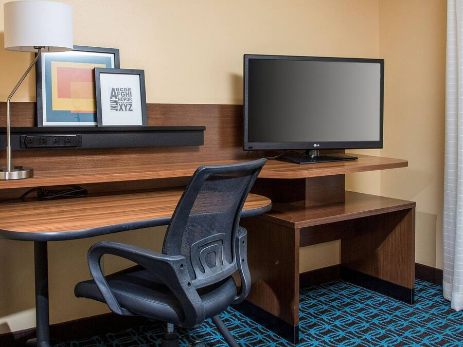 Habitación de hotel con un escritorio de madera, una silla de oficina negra, un televisor de pantalla plana, una lámpara y obras de arte enmarcadas en una estantería.