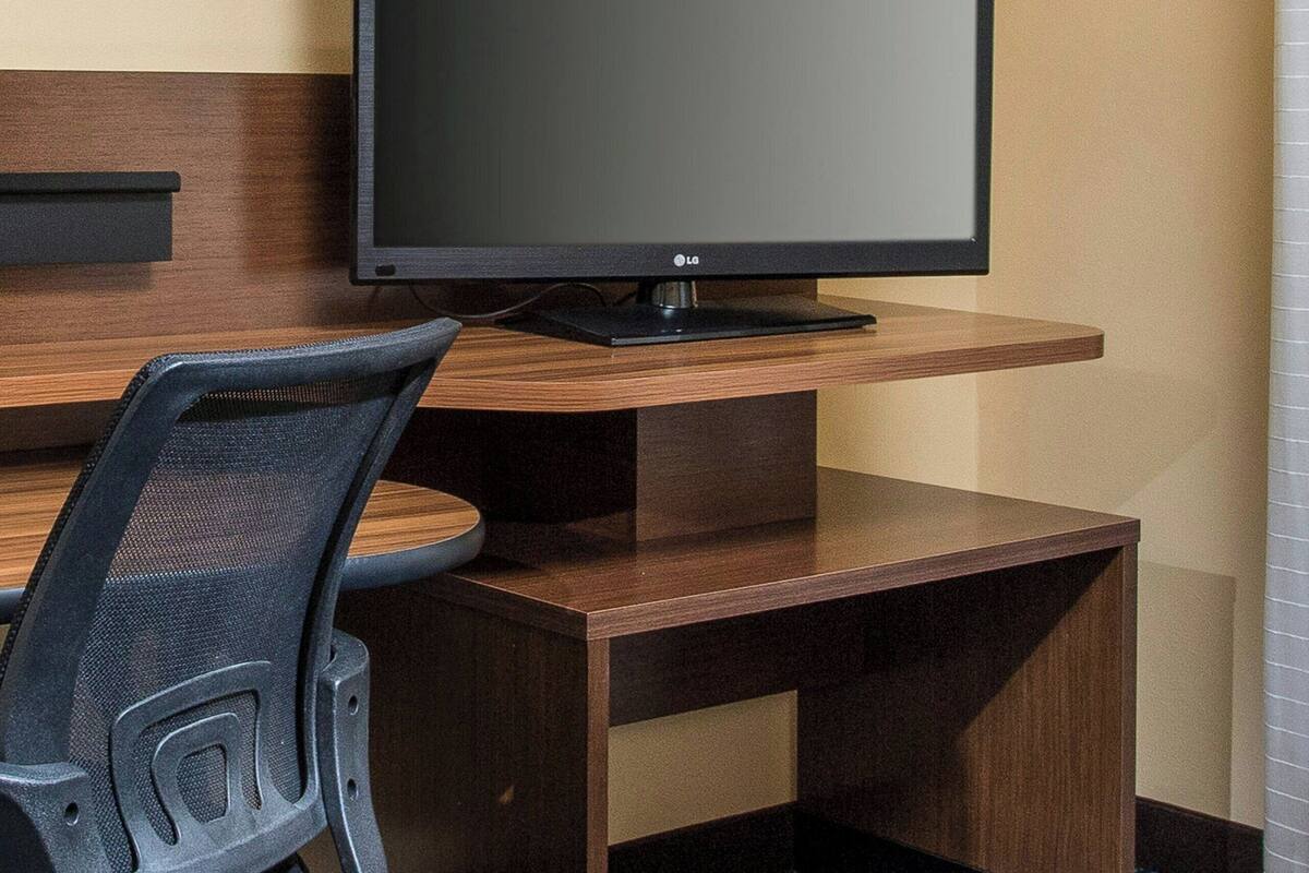 A hotel room with a wooden desk, a black office chair, a flat-screen TV, and a patterned carpet.