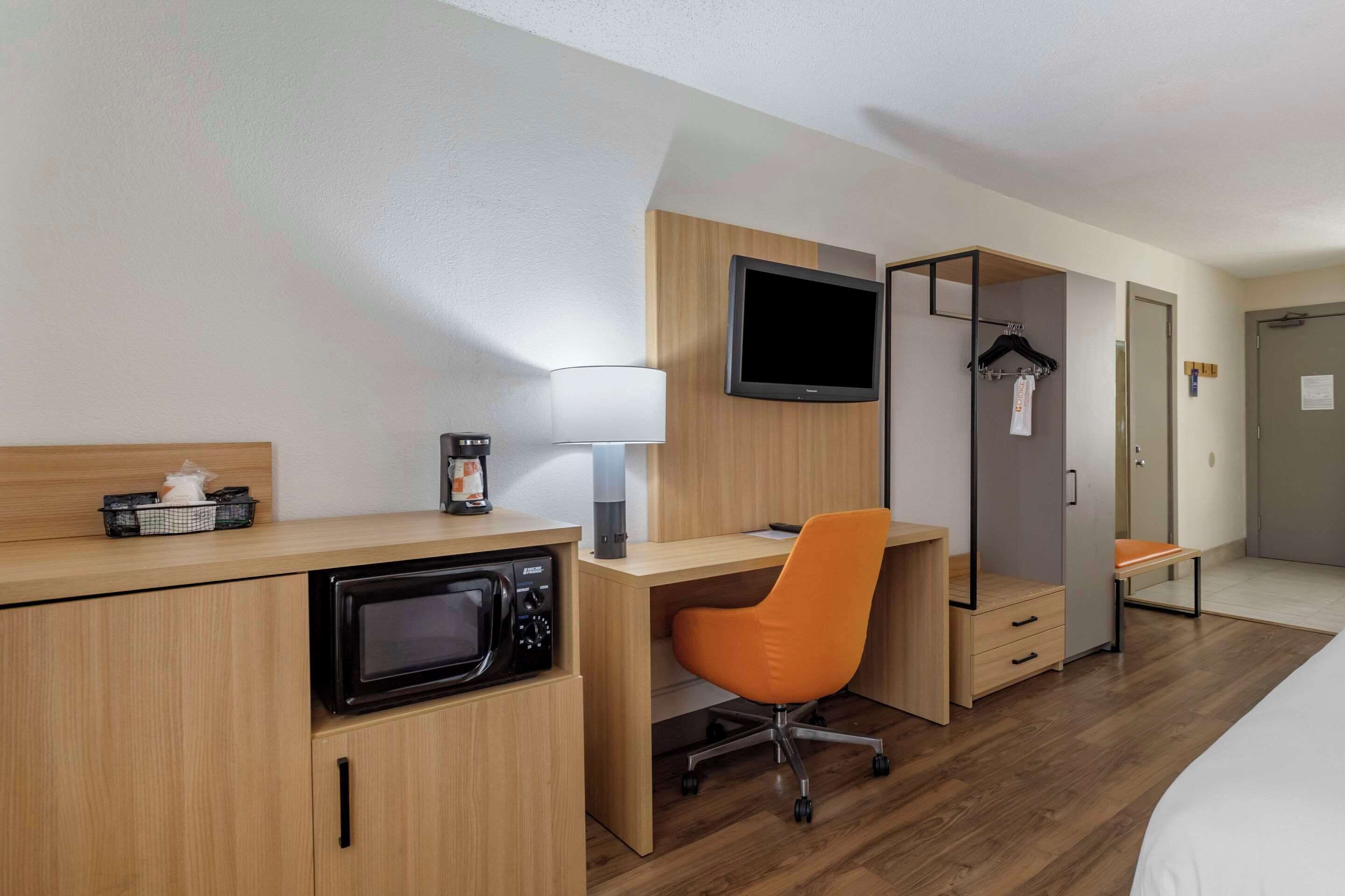 Habitación de hotel con escritorio de madera, microondas, televisión, una silla naranja, cama y armario.