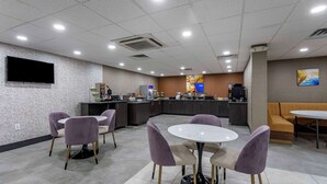 Un moderno vestíbulo de hotel con una mesa redonda central y sillas moradas, un televisor de pantalla plana en la pared y una zona de cocina con microondas y refrigerador.
