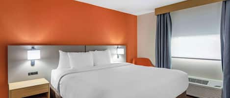 Quarto de hotel com uma cama grande, uma mesa de cabeceira, uma cadeira, uma luminária na parede e uma janela com cortinas.