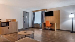 Una habitación de hotel moderna con suelo de madera, un sofá, una pequeña mesa redonda, un escritorio con una silla naranja, un televisor de pantalla plana montado en la pared y una lámpara.