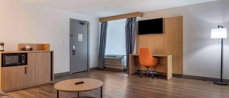 A modern hotel room with a wooden floor, a sofa, a small round table, a desk with an orange chair, a flat-screen TV mounted on the wall, and a lamp.