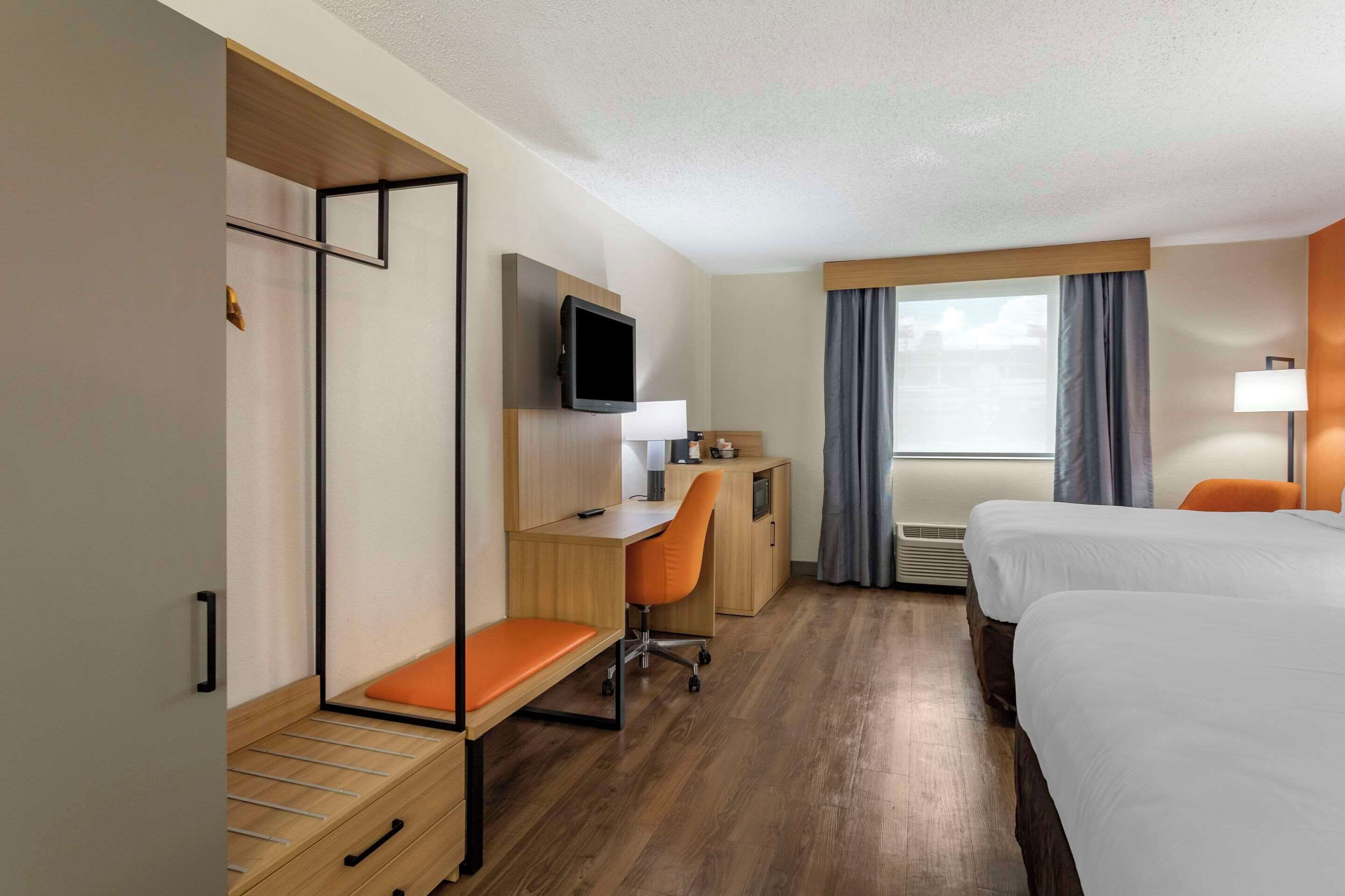 A hotel room with a bed, a desk with a chair, a TV, a window with curtains, and a bedside lamp.