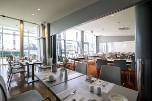 A spacious dining area with multiple tables and chairs, large windows offering views of the city, and a modern interior design. - Nordsee Hotel Bremerhaven Fischereihafen (Bremerhaven)