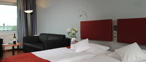 A hotel room with a bed, a black leather chair, a small table with a bottle and glass, a vase with flowers, and a red headboard.
