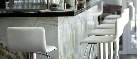 Zona de bar con mostrador de mármol, taburetes blancos y una variedad de botellas y vasos en estanterías.