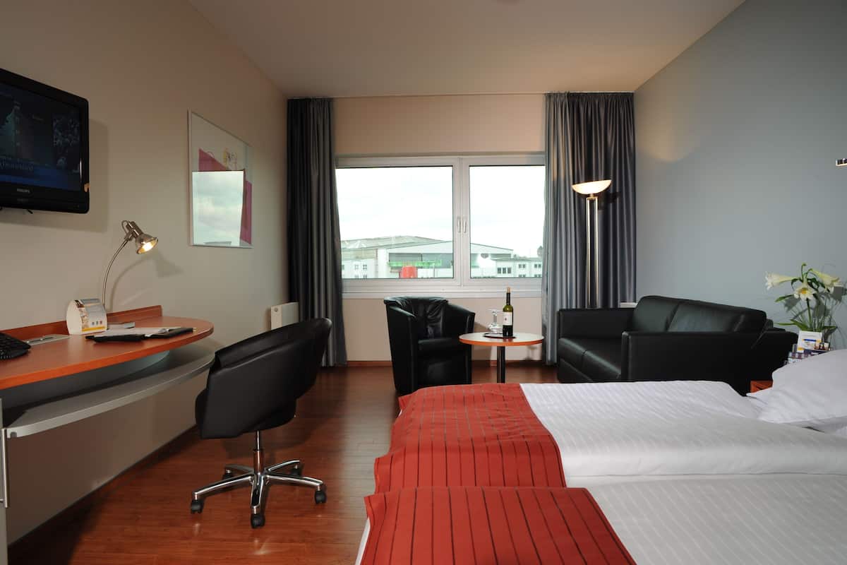 A hotel room with a bed, a desk, two chairs, a small table with a bottle, a lamp, a TV, and a window with curtains.