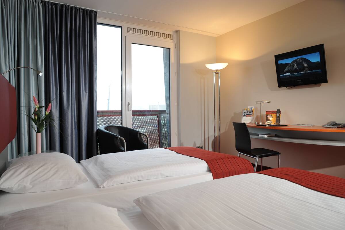A hotel room with two beds, a desk, a chair, a TV, and a window with curtains.