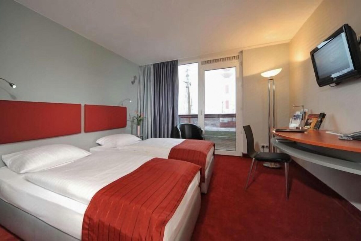 Une chambre d’hôtel avec un grand lit, un bureau, une chaise, une télévision et une fenêtre avec des rideaux.