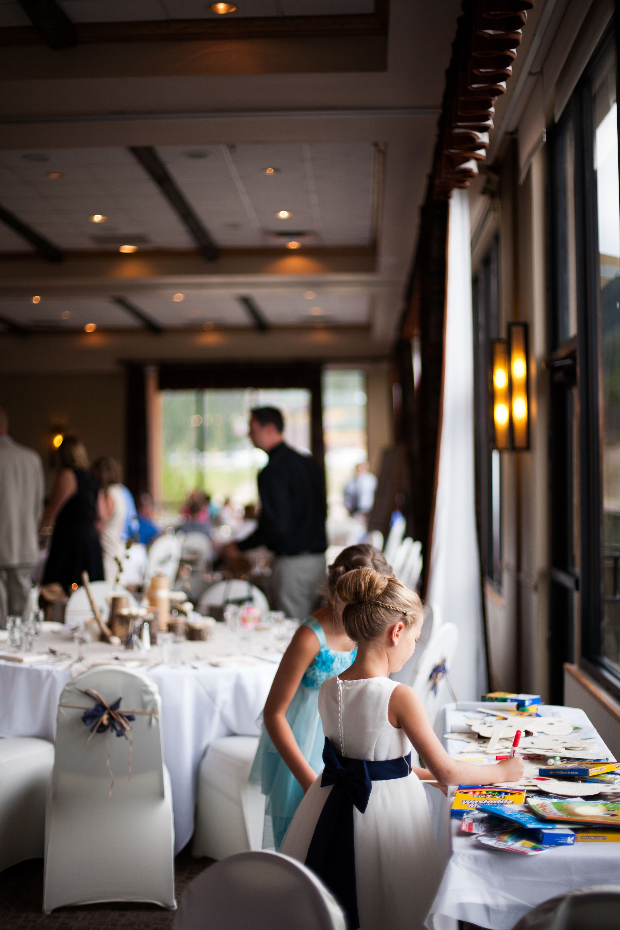 A formal dining area with tables set for a meal, a young girl painting at a table, and a man in the background.