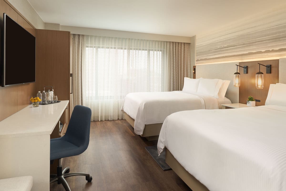 A hotel room with a flat-screen TV, a desk with a chair, two beds, a large window with sheer curtains, and wall-mounted lamps.