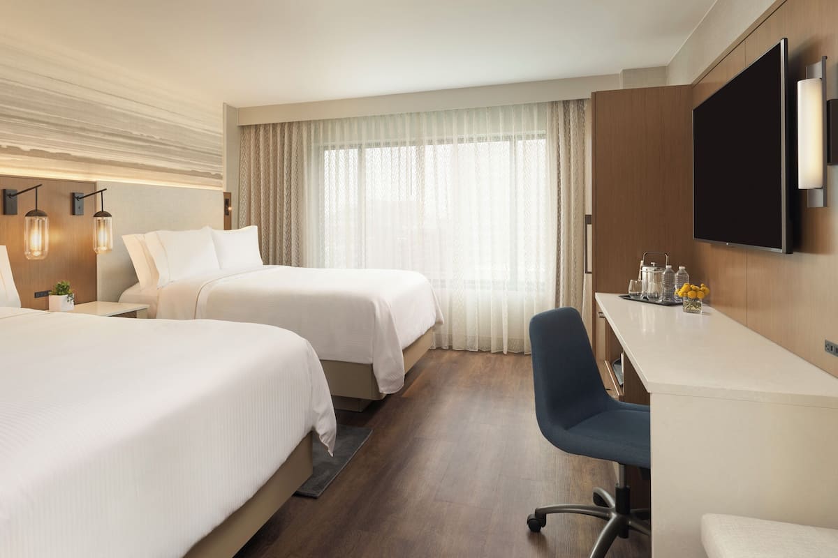 A hotel room with two beds, a desk, a chair, a TV, and a window with curtains.