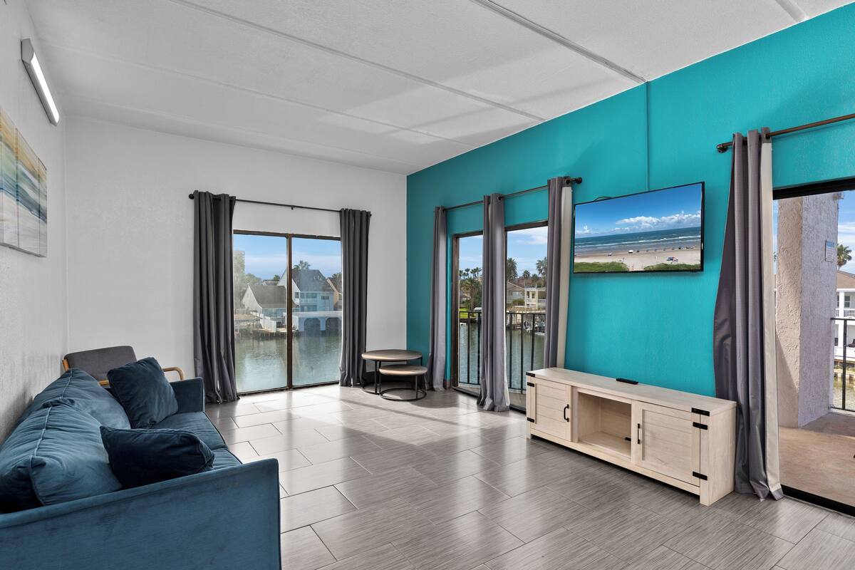 A modern living room with a teal accent wall, a sofa, a TV, and a view of the beach.