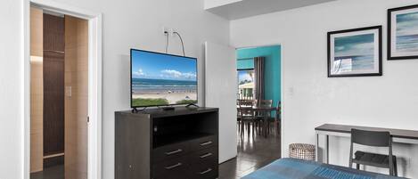 A modern hotel room with a large bed, a flat-screen TV on a wooden cabinet, a desk with a chair, and a dining area with a table and chairs.