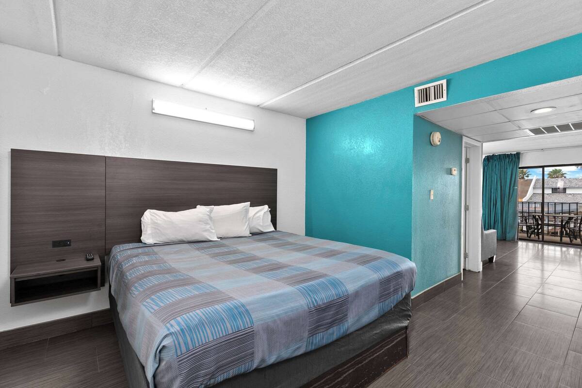 A hotel room with a large bed, a nightstand, a built-in shelf, a window with curtains, and a view of the outside balcony.