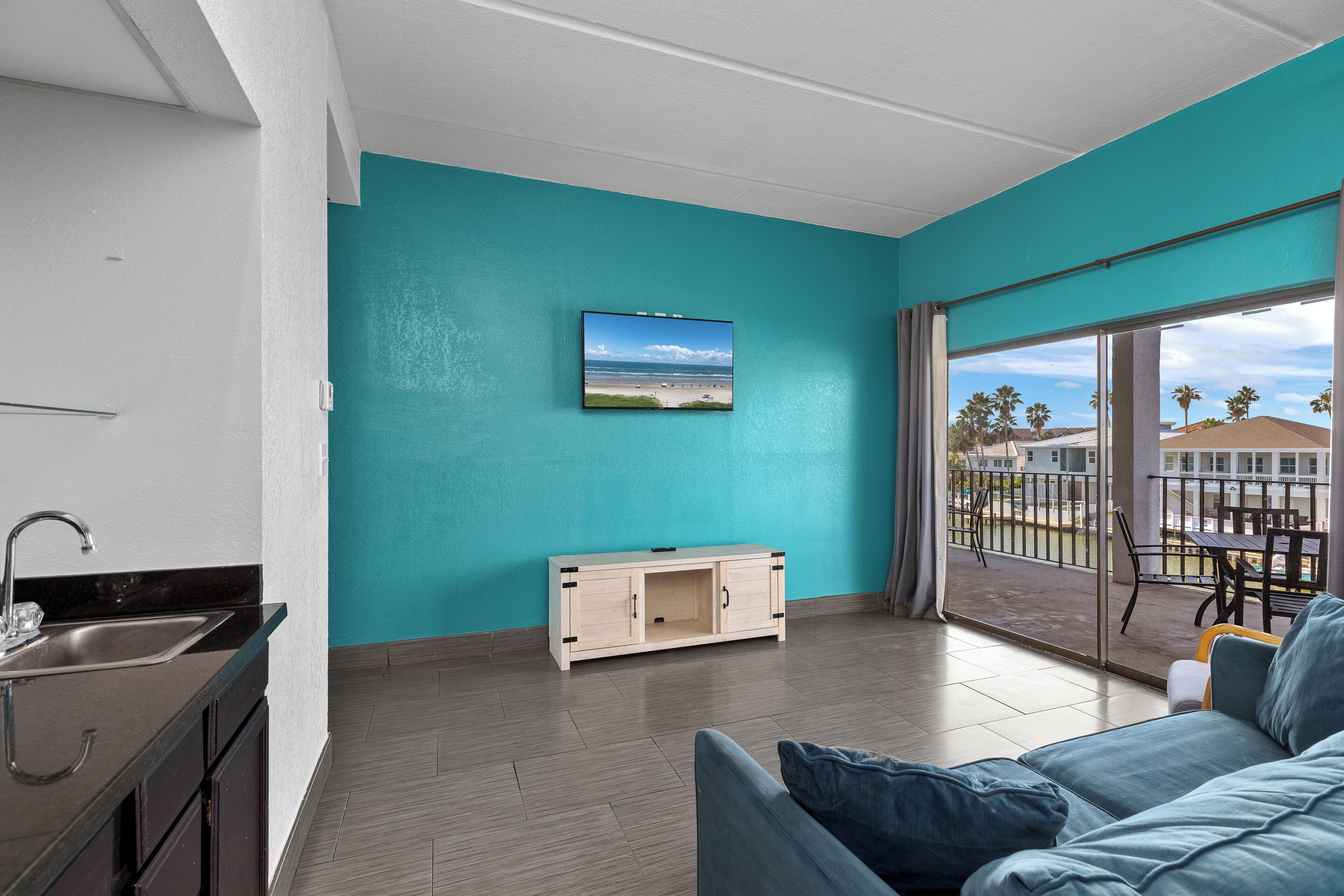 Ein modernes Wohnzimmer mit einer türkisfarbenen Wand, einem Flachbildfernseher, einer Couch und einem Balkon mit Blick auf Palmen und einen Strand.
