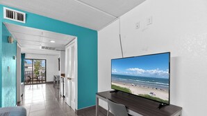 Habitación de hotel con una televisión mostrando una vista de la playa, una zona de comedor con mesa y sillas, y una puerta que da a un balcón con vistas a la playa.