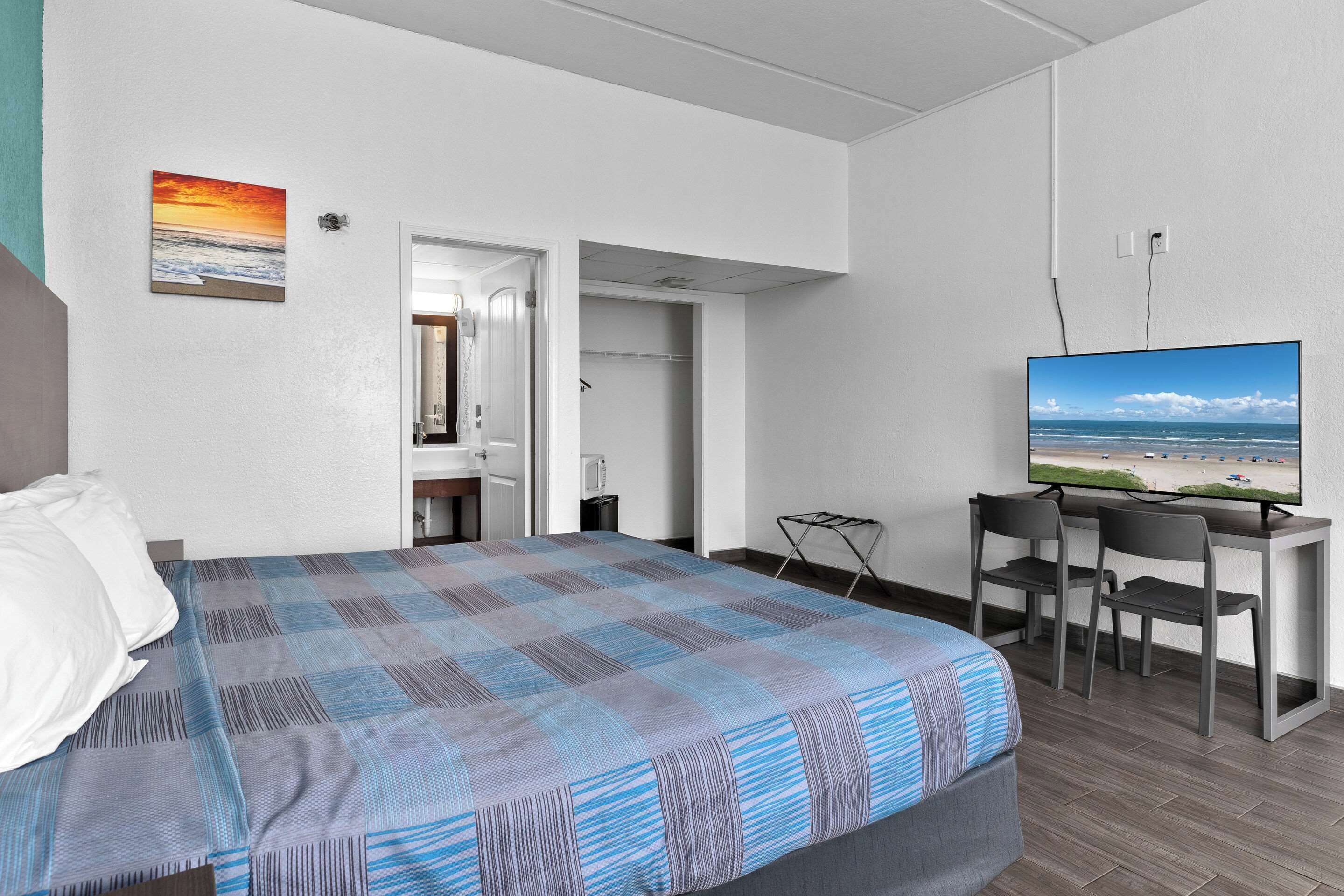 Quarto de hotel com uma cama, uma escrivaninha com duas cadeiras, uma televisão e uma pintura de um pôr do sol.