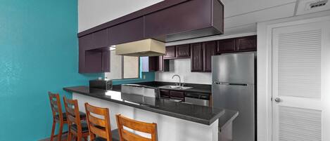 A modern kitchen with a central island, bar stools, dark wooden cabinets, and a stainless steel refrigerator.