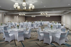 Een ruime balzaal met ronde tafels, witte stoelen en kroonluchters.