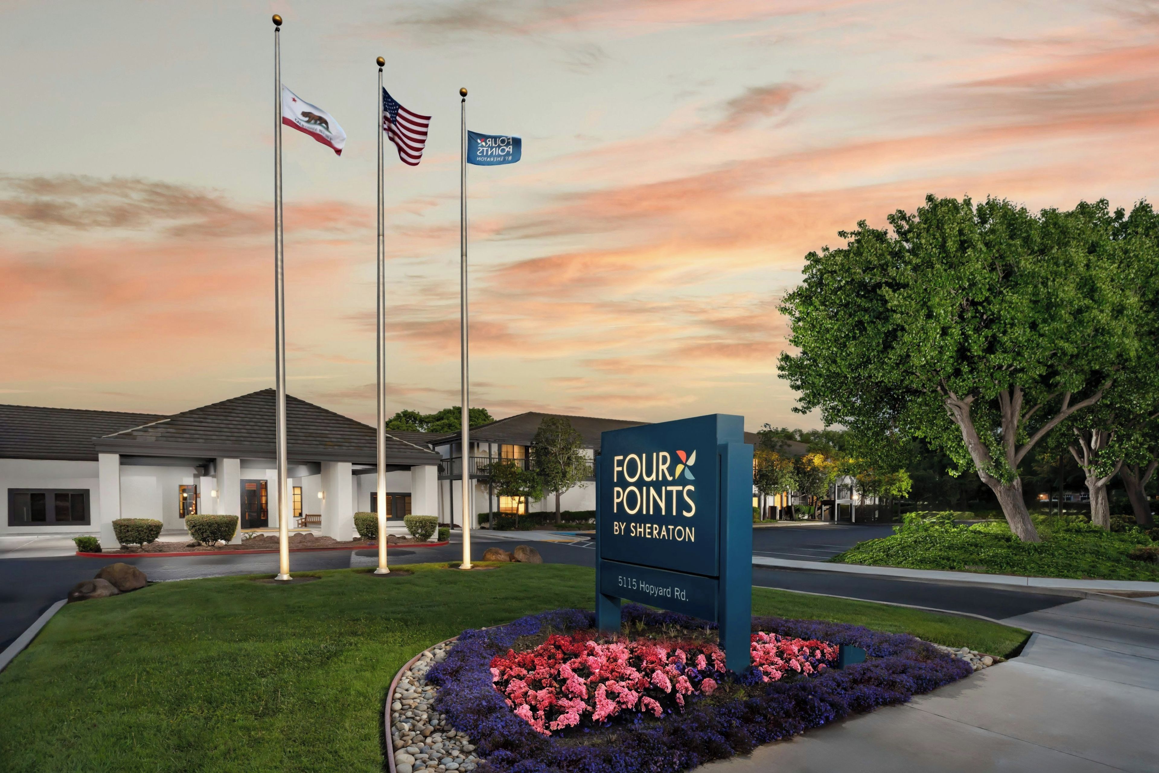 A hotel entrance with a sign, flags, and a landscaped area.