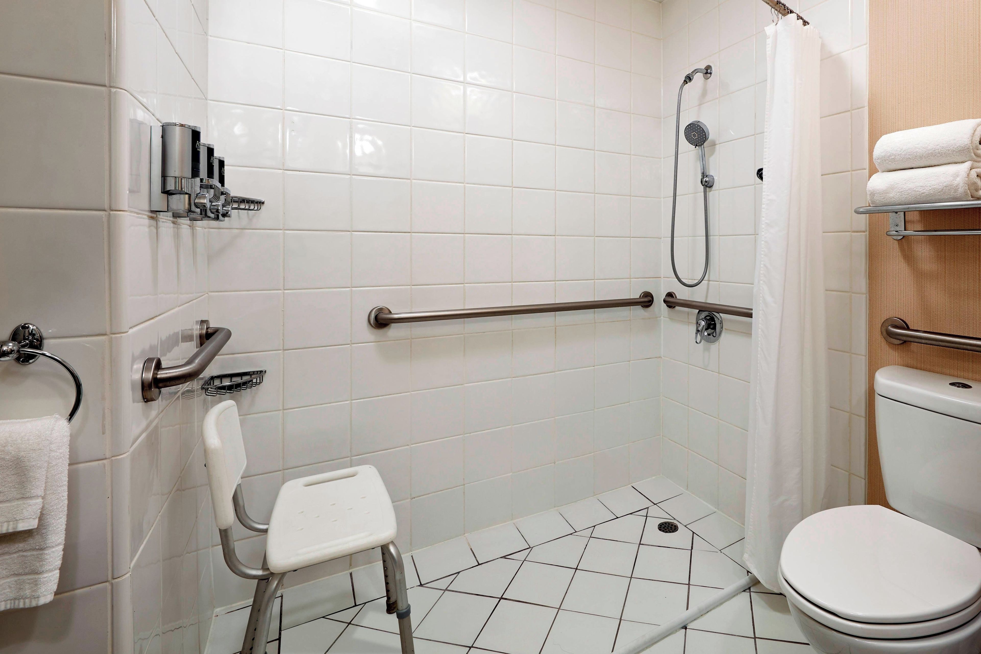 A wheelchair-accessible shower area with a grab bar, a white toilet, and a small white chair.