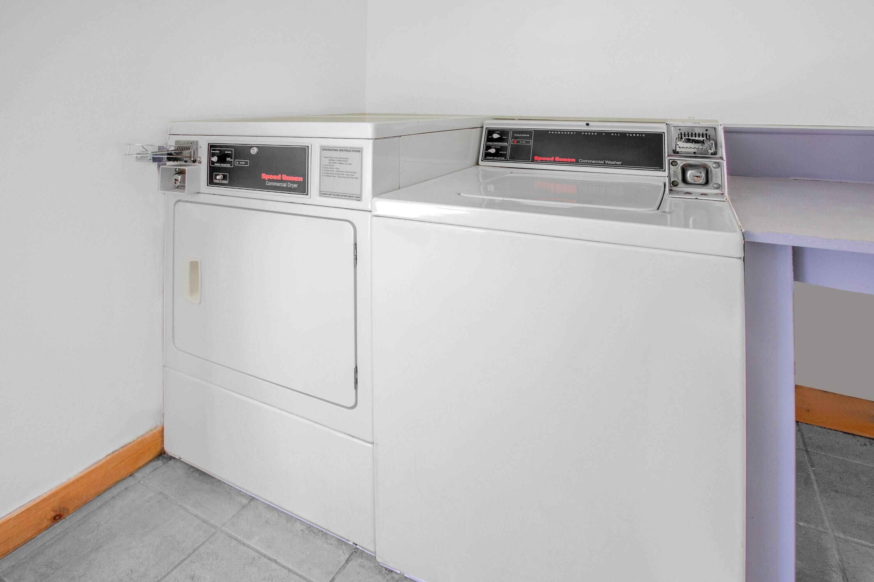 A white commercial washer with control panels and a door, located in a corner of a room with tiled flooring and a wooden baseboard.