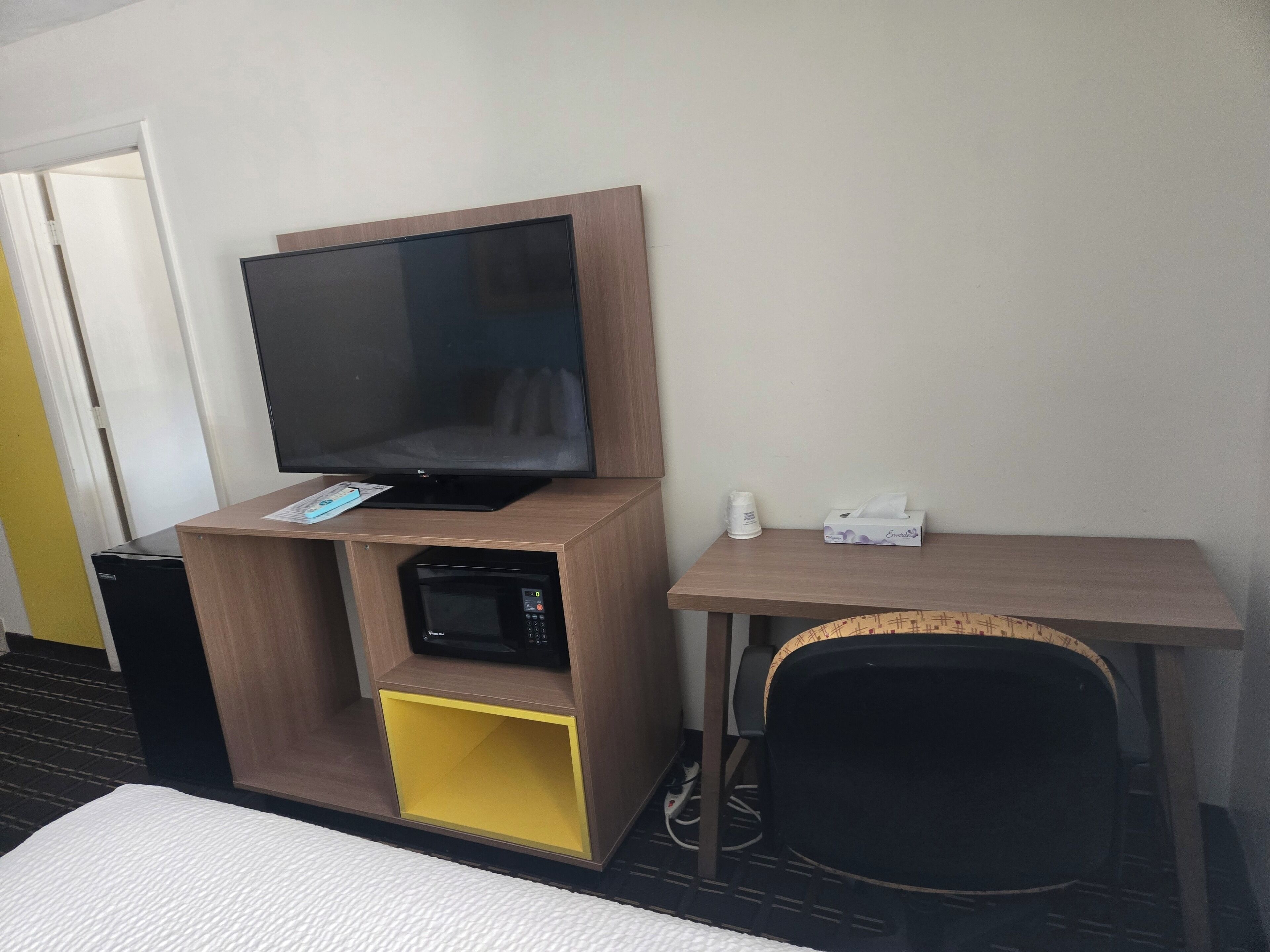 Habitación de hotel con televisor de pantalla plana sobre un soporte de madera, microondas, una mesita con una caja de pañuelos y una silla de mimbre.