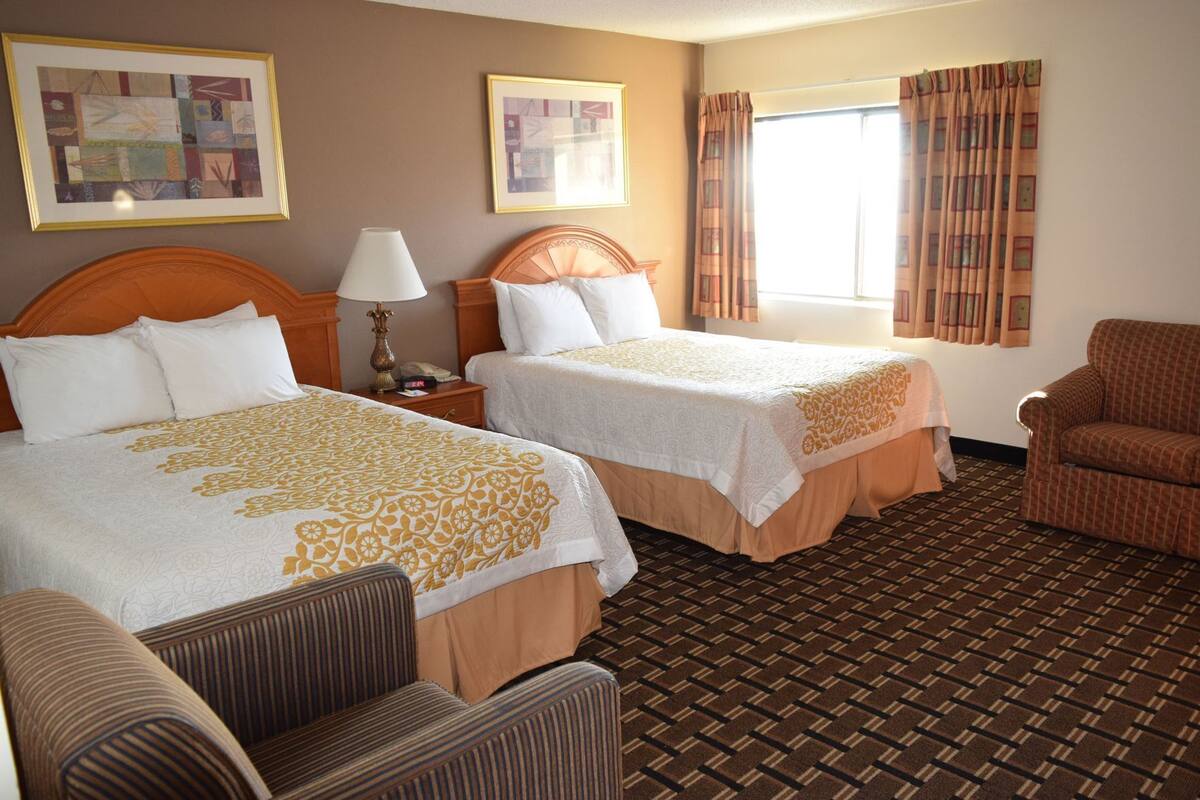 A hotel room with two beds, a chair, a lamp, and a window with curtains.