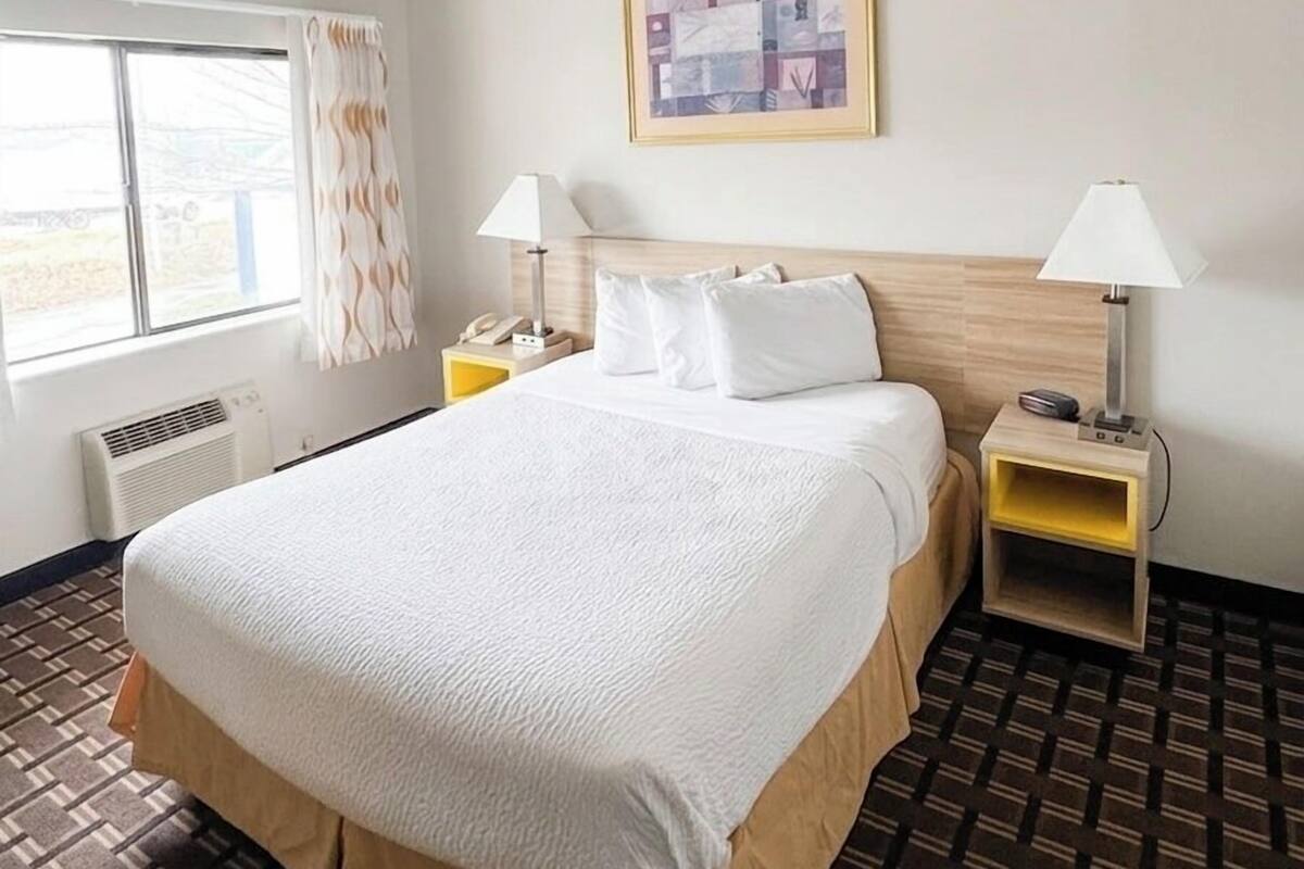 A hotel room with a large bed, two bedside tables, a window with curtains, a radiator, and a framed picture on the wall.