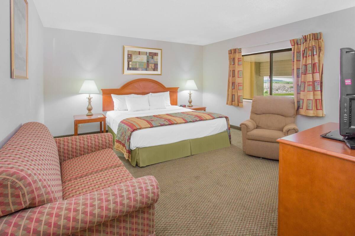 A hotel room with a bed, two bedside lamps, a chair, a desk with a television, and a window with curtains.
