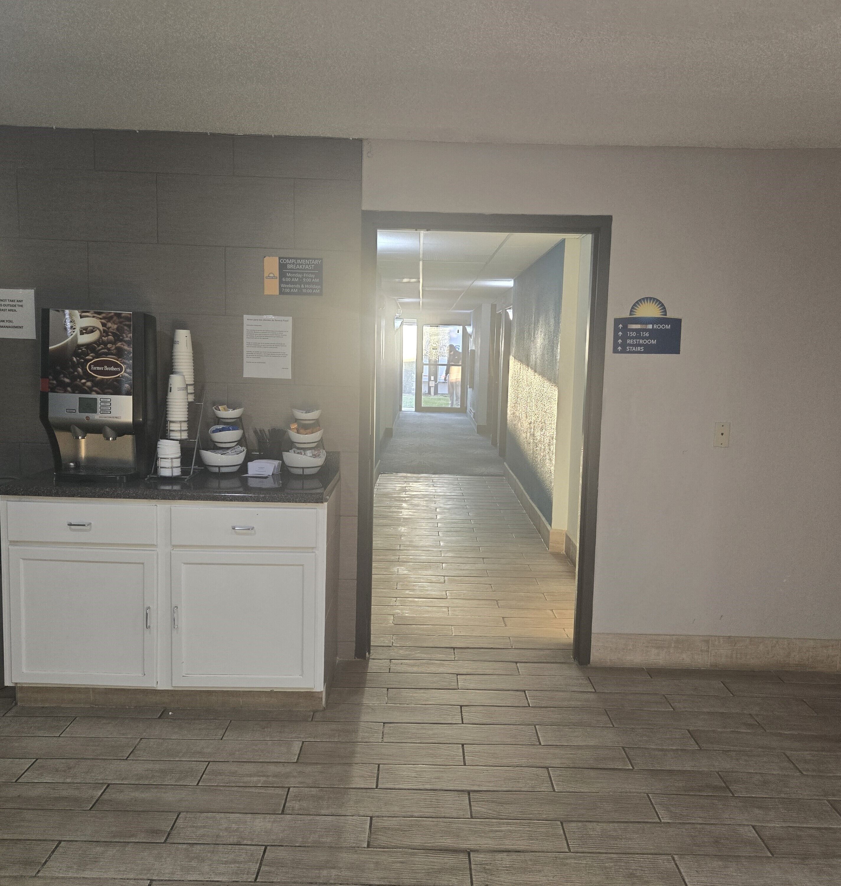 A hotel corridor with a coffee machine, complimentary breakfast sign, and directional signs for room, restroom, and stairs.
