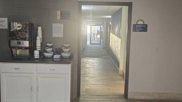 A hotel corridor with a coffee machine, complimentary breakfast sign, and directional signs for room, restroom, and stairs.