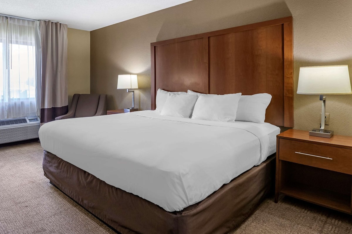A hotel room with a large bed, a wooden headboard, a bedside table with a lamp, a chair, and a window with curtains.