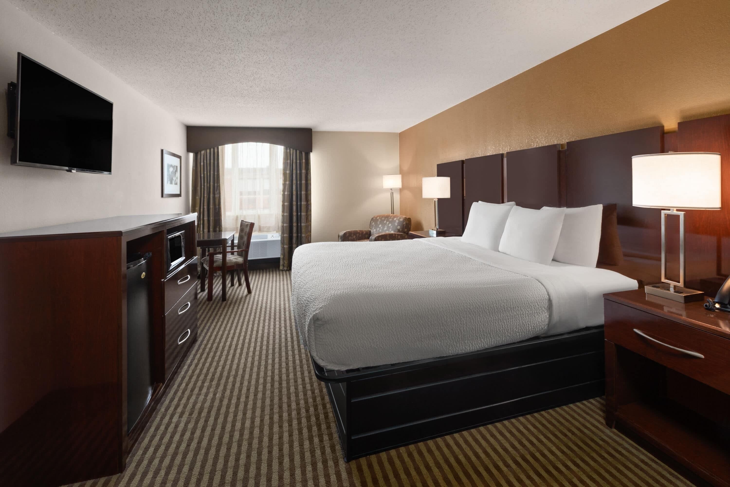 A hotel room with a large bed, a desk, a chair, a TV mounted on the wall, and a window with curtains.