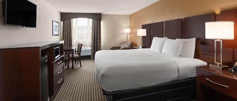 A hotel room with a large bed, a desk, a chair, a TV mounted on the wall, and a window with curtains.