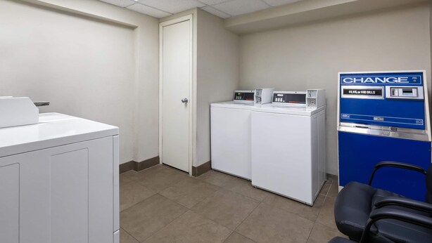 A laundry room with two washing machines, a change machine, and a chair. - Baymont by Wyndham Festus (Festus)