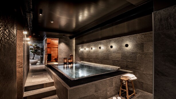 A modern spa area with a heated stone bath, a wooden stool, and a small plant.