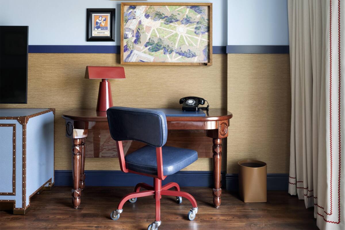 A hotel room with a wooden desk, a red office chair, a framed artwork, a telephone, and a waste bin.